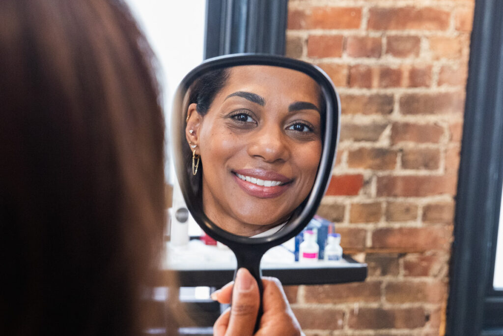 A woman is holding up a mirror and smiling after seeing the results from her acne scar treatment in Newburgh, NY.